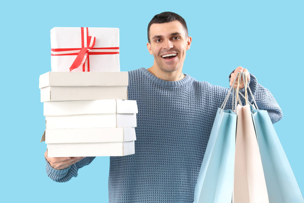 Schöner junger glücklicher Mann mit Geschenkschachteln und Einkaufstaschen auf blauem Hintergrund - Foto, Bild
