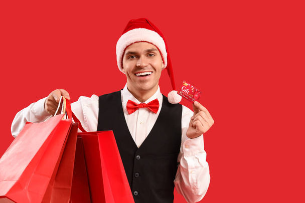 Schöner junger glücklicher Mann mit Weihnachtsmütze mit Geschenkkarte und Einkaufstaschen auf rotem Hintergrund - Foto, Bild