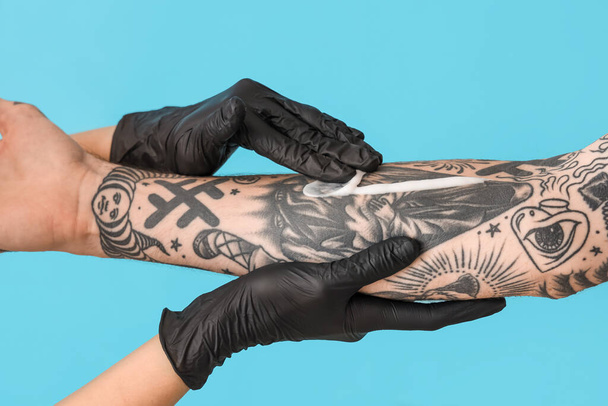 Maestro del tatuaje en guantes aplicando crema en el brazo del hombre con tatuajes sobre fondo azul, primer plano - Foto, Imagen