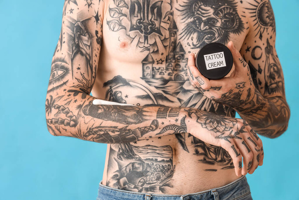 Homme tatoué tenant un pot de crème de tatouage sur fond bleu, gros plan - Photo, image