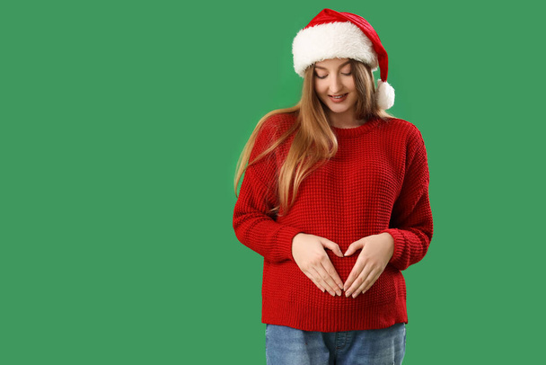 Hermosa joven embarazada en Santa sombrero haciendo corazón con sus manos sobre fondo verde - Foto, Imagen