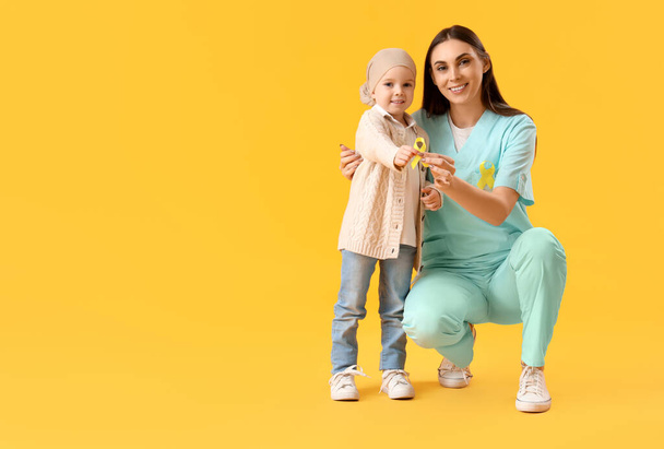Kleines Mädchen nach der Chemotherapie mit Krankenschwester und Schleife auf gelbem Hintergrund. Konzept zur Sensibilisierung für Kinderkrebs - Foto, Bild