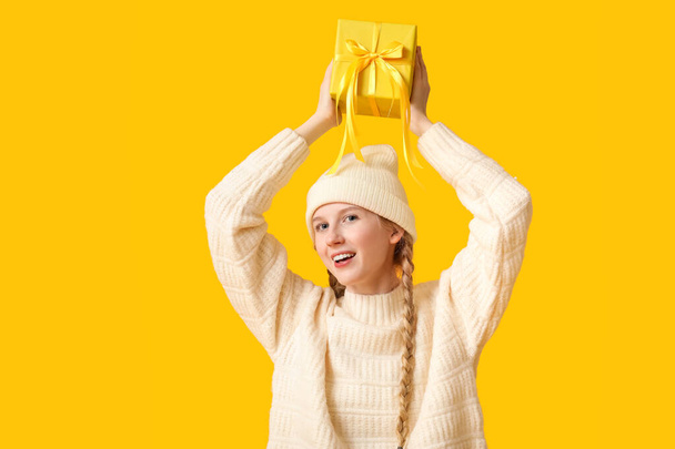 Beautiful young woman in warm winter clothes with gift box on yellow background - Photo, Image