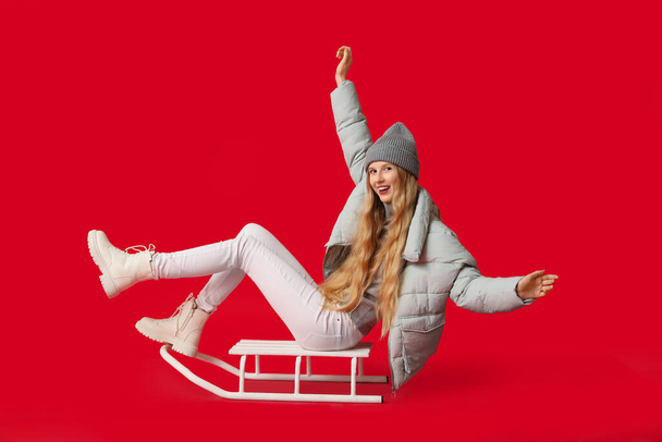 Beautiful young happy woman in warm winter clothes with sledge on red background - Photo, Image