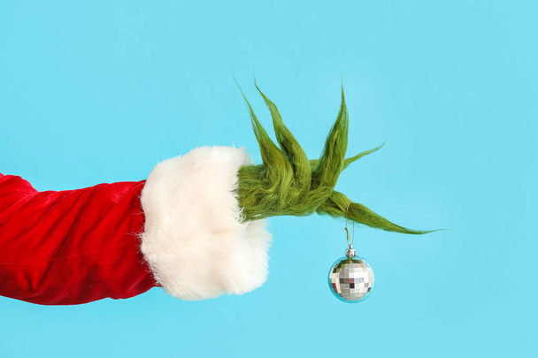 Grüne haarige Hand der Kreatur im Weihnachtsmannkostüm mit Weihnachtskugel auf blauem Hintergrund - Foto, Bild