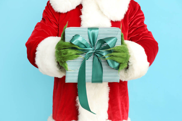 Green hairy creature in Santa costume with gift box on blue background, closeup - Photo, Image