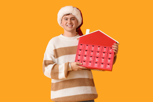 Happy young man in Santa hat with Christmas advent calendar on yellow background - Photo, Image