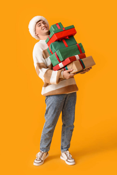 Happy young man in Santa hat with Christmas gifts on yellow background - Photo, Image