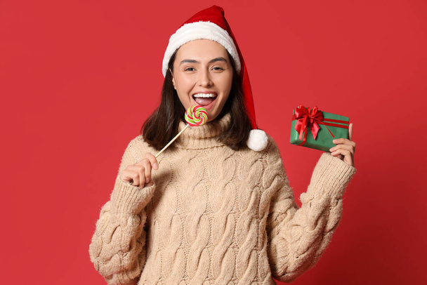 Happy young woman in Santa hat with lollipop and Christmas gift on red background - Photo, Image