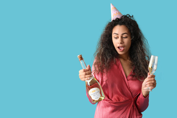美しい若いアフリカ系アメリカ人女性 パーティーハットで ガラスとシャンパンボトル 青い背景で誕生日を祝います - 写真・画像