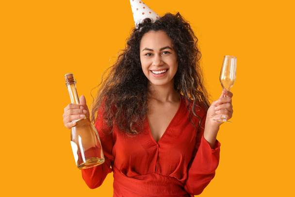 黄色の背景で誕生日を祝うシャンパンとガラスとボトルでパーティー帽子で美しい若いアフリカ系アメリカ人女性 - 写真・画像