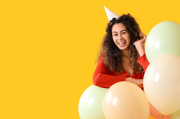 Krásná mladá afroamerická žena ve stranickém klobouku se vzduchovými balónky slaví narozeniny na žlutém pozadí - Fotografie, Obrázek