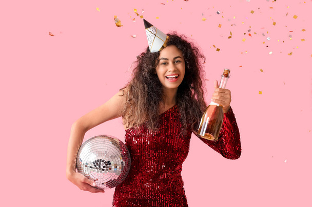 美しい若いアフリカ系アメリカ人女性,ディスコボールとピンクの背景で誕生日を祝うシャンパンのボトルでパーティー帽子 - 写真・画像