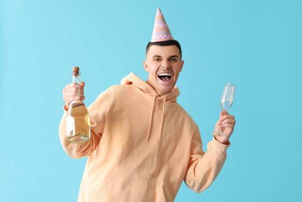 Mladý muž ve stranickém klobouku se sklem a lahví šampaňského na modrém pozadí - Fotografie, Obrázek