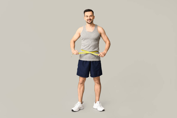 Knappe sportieve jongeman die zijn middel meet op een grijze achtergrond. Gewichtsverlies concept - Foto, afbeelding