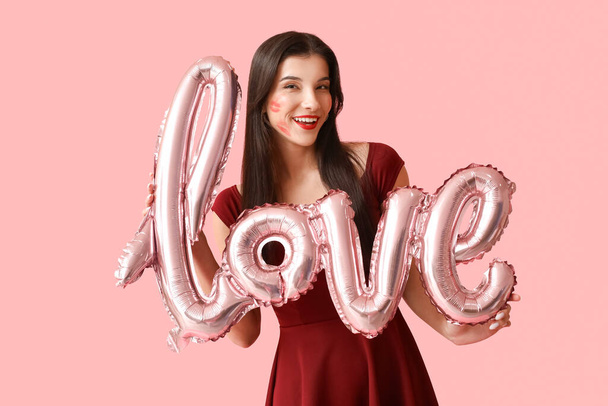 Šťastná mladá žena s polibky značky na tváři a vzduchové balónky ve tvaru slova LOVE na růžovém pozadí - Fotografie, Obrázek