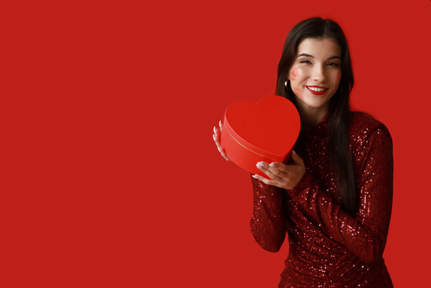 Счастливая молодая женщина со следами поцелуев на лице и подарочной коробкой на красном фоне - Фото, изображение