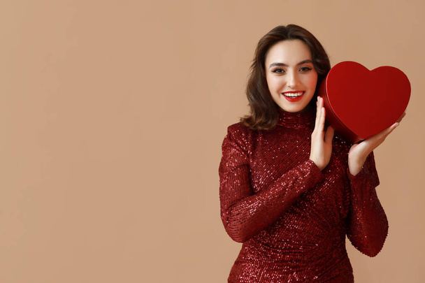 Щаслива молода жінка з подарунковою коробкою у формі серця на коричневому фоні - Фото, зображення
