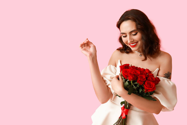Beautiful young woman with bouquet of red roses on pink background - Photo, Image