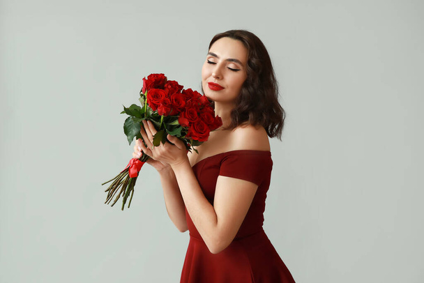Όμορφη νεαρή γυναίκα με μπουκέτο κόκκινα τριαντάφυλλα σε γκρι φόντο - Φωτογραφία, εικόνα