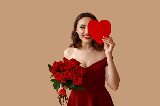 Mujer joven feliz con ramo de rosas y corazón de papel rojo sobre fondo marrón - Foto, imagen