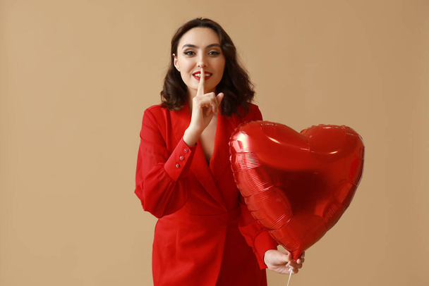 mooie jonge vrouw met hartvormige luchtballon tonen stilte gebaar op bruine achtergrond - Foto, afbeelding