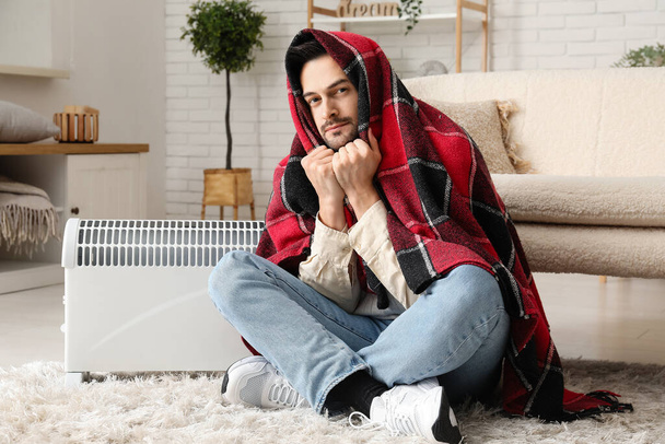 Sick young man with plaid warming near radiator at home - Photo, Image