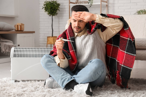 Sick young man with plaid and thermometer warming near radiator at home - Photo, Image