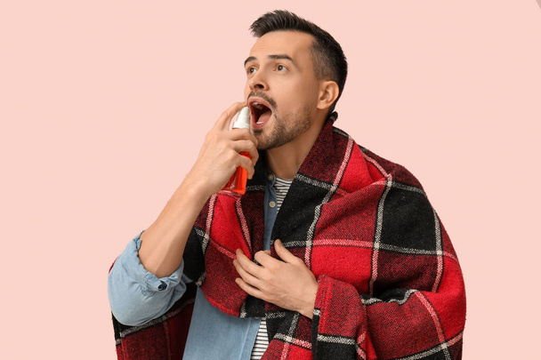 Sick young man with plaid using spray for sore throat on pink background - Photo, Image