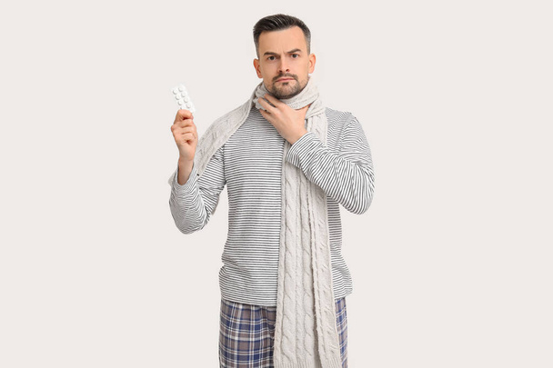 Nemocný mladý muž v šále s pilulkami na lehkém pozadí - Fotografie, Obrázek