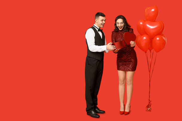 Αγαπώντας νεαρό ζευγάρι με μπαλόνια σε σχήμα καρδιάς και κουτί δώρου σε κόκκινο φόντο. Εορτασμός της ημέρας του Αγίου Βαλεντίνου - Φωτογραφία, εικόνα