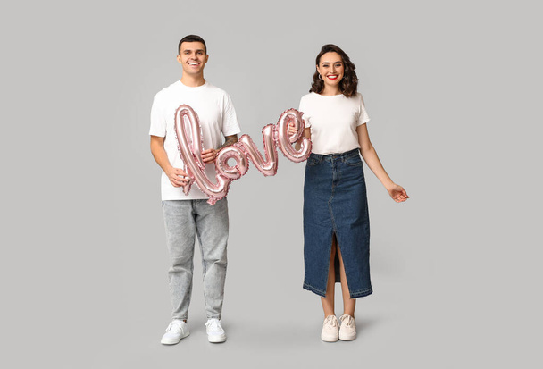 Αγαπώντας νεαρό ζευγάρι με μπαλόνι σε σχήμα λέξης ΑΓΑΠΗ σε γκρι φόντο. Εορτασμός της ημέρας του Αγίου Βαλεντίνου - Φωτογραφία, εικόνα