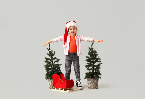 Schattig klein meisje in Santa hoed met kerstbomen en slee op lichte achtergrond - Foto, afbeelding