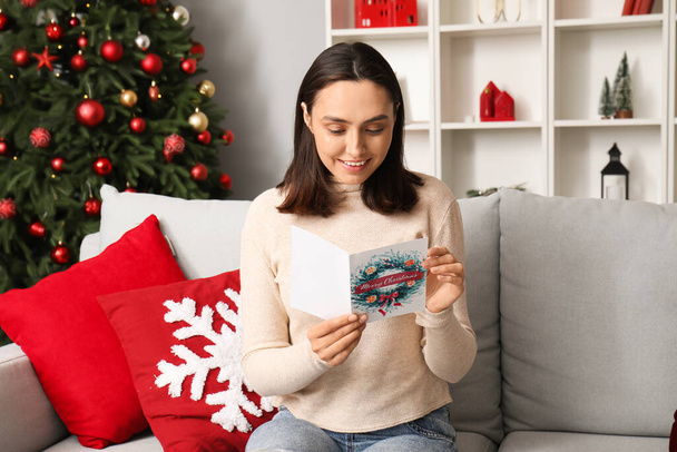 Glückliche junge Frau liest Weihnachtsgrußkarte, während sie auf dem Sofa im festlichen Wohnzimmer sitzt - Foto, Bild