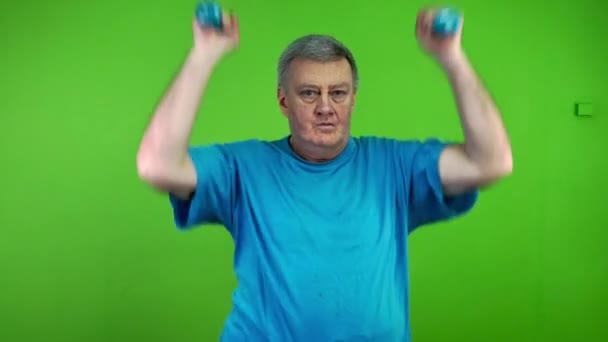 Az idősebb férfi súlyzózik, naponta edz, fel-le mozgatja a kezét. Idős ember sportol vagy testmozgást végez az egészség fenntartása érdekében. Zöld vászon. Kroma kulcs. - Felvétel, videó