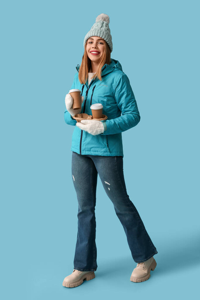 Piękna młoda szczęśliwa kobieta w ciepłych zimowych ubraniach z filiżankami kawy na niebieskim tle - Zdjęcie, obraz