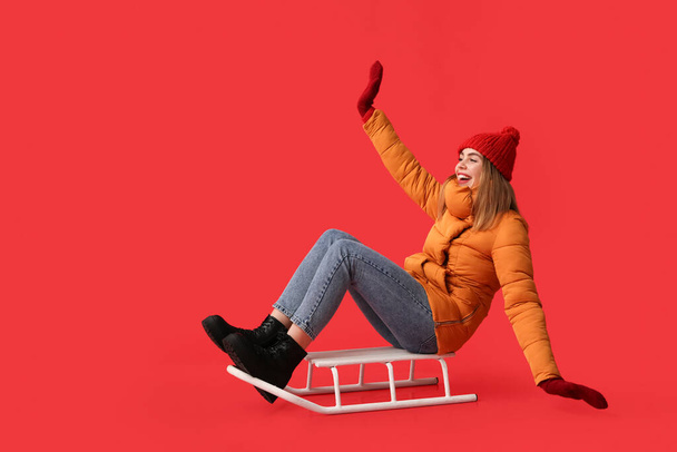 Όμορφη νεαρή ευτυχισμένη γυναίκα σε ζεστά χειμωνιάτικα ρούχα με έλκηθρο κάθεται σε κόκκινο φόντο - Φωτογραφία, εικόνα