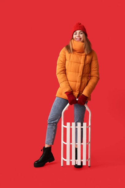 Όμορφη νεαρή ευτυχισμένη γυναίκα σε ζεστά χειμωνιάτικα ρούχα με έλκηθρο σε κόκκινο φόντο - Φωτογραφία, εικόνα