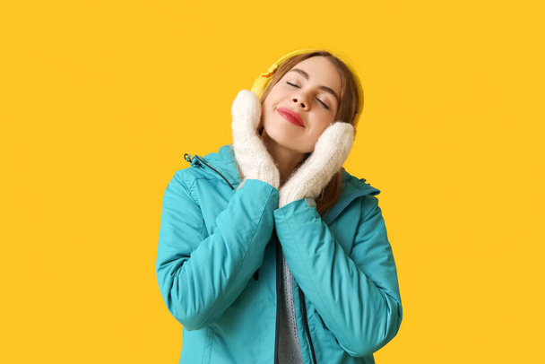 Sarı arka planda kulaklıkları olan sıcak kış kıyafetleri içinde güzel, genç bir kadın. - Fotoğraf, Görsel