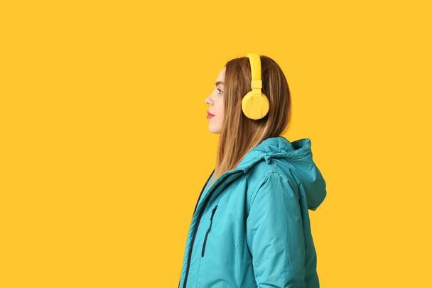 Beautiful young woman in warm winter clothes with headphones on yellow background - Photo, Image