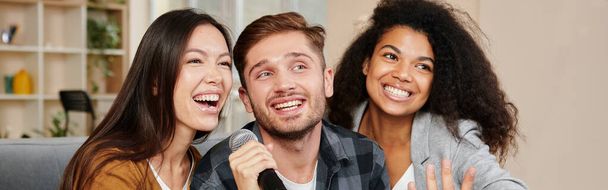 Karaokelaulajia. Kolme hymyilevää ystävää, jotka näyttävät onnellisilta pelatessaan karaokea kotona, laulaen mikrofonilla, istuen sohvalla modernissa asunnossa. Karaokejuhlat, ystävyyden käsite - Valokuva, kuva
