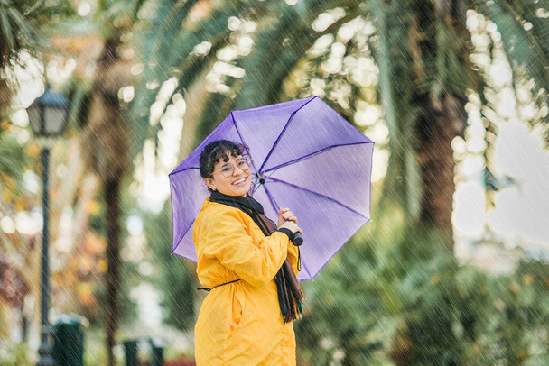 Mujer amigable venezolana que usa protección de abrigo amarillo para el pronóstico de lluvia. Estilo de vida persona alegre en ambiente lluvioso con espacio de copia - Foto, Imagen