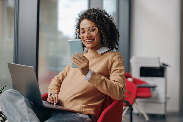 Stylowa kobieta biznesu jest korzystanie z telefonu podczas pracy na laptopie w nowoczesnym biurze w pobliżu okna - Zdjęcie, obraz