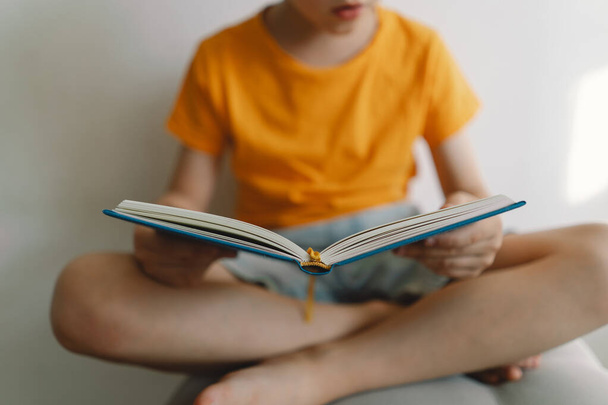 Ένα χαριτωμένο αγόρι που φοράει πορτοκαλί μπλουζάκι κάθεται πάνω σε ένα μαλακό κοτόμανο διαβάζοντας ένα βιβλίο. Αντιγραφή χώρου. Πραγματικοί άνθρωποι και τρόπος ζωής - Φωτογραφία, εικόνα