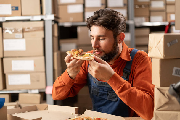 Valkoihoinen mies työvaatteissa, varastotyöntekijä syö lihapizzaa pikaruoasta lounaaksi istuessaan pöydässä.. - Valokuva, kuva