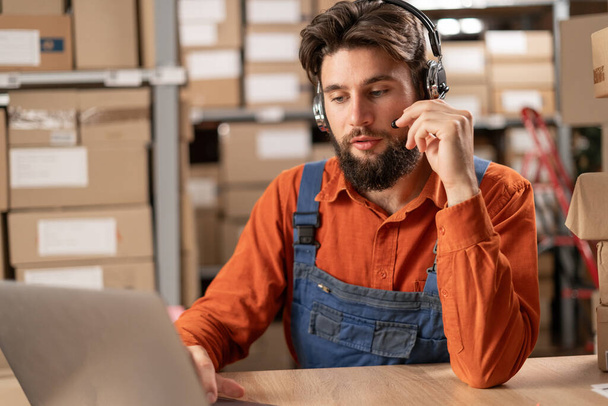A technical support operator at a delivery warehouse works at a laptop using a headset. Smiling handsome man talking to a client. - Photo, Image