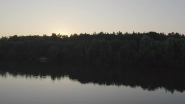 Ten spokojny materiał rejestruje zachód słońca za gęstym lasem, jego gasnące światło rzuca spokojny blask nad spokojnym jeziorem. Odbicie drzew na powierzchni wody zwiększa poczucie - Materiał filmowy, wideo