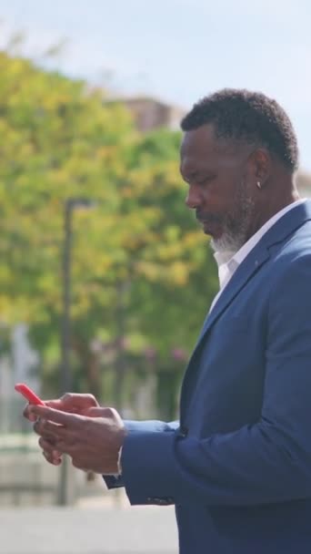 Video sivusuunnassa näkymä kypsä afrikkalainen liikemies lähettää viestin puhelimitse ulkona - Materiaali, video