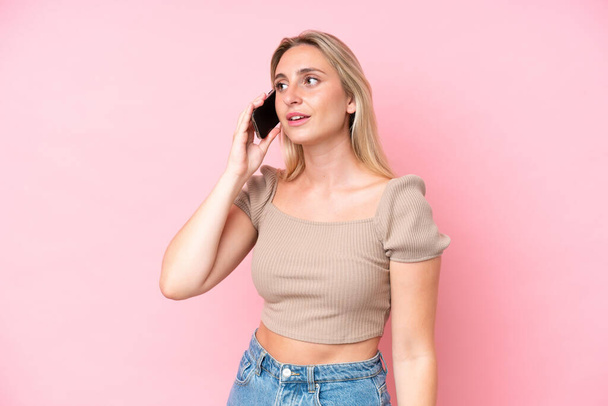 Jonge blanke vrouw geïsoleerd op roze achtergrond houden van een gesprek met de mobiele telefoon - Foto, afbeelding