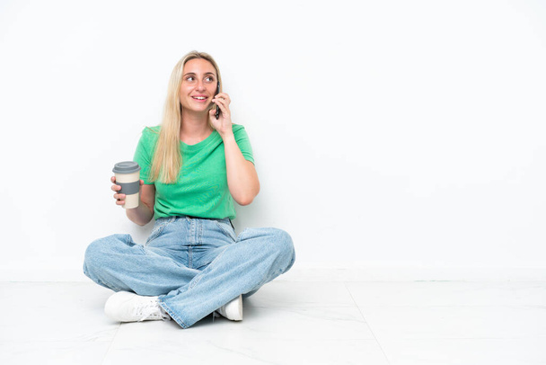 Mladá běloška sedící na podlaze izolované na bílém pozadí držící kávu odnést a mobilní - Fotografie, Obrázek